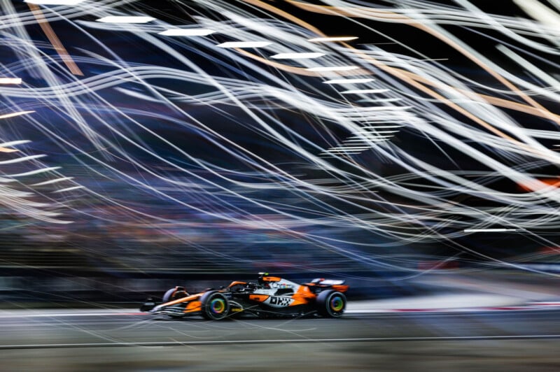 A Formula 1 race car speeds along a track at night, with bright, streaking lights creating dynamic motion trails above and behind the vehicle. The background is blurred, emphasizing the car’s rapid movement.