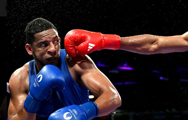 A boxer wearing blue gloves ducks as a red-gloved punch narrowly misses his face in a dramatic boxing match, with droplets of sweat flying in the air under bright lights.