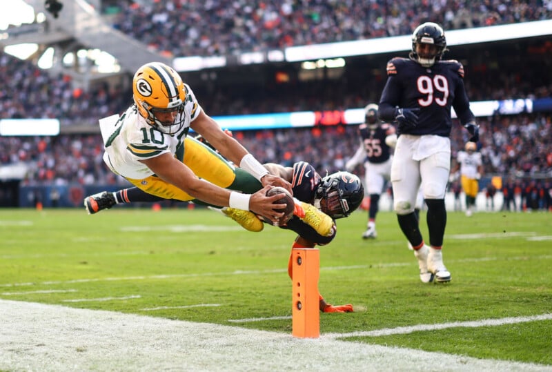 A Green Bay Packers player dives toward the end zone, reaching the football over the goal line as a Chicago Bears defender tries to stop him, with another Bears player watching nearby during a game.