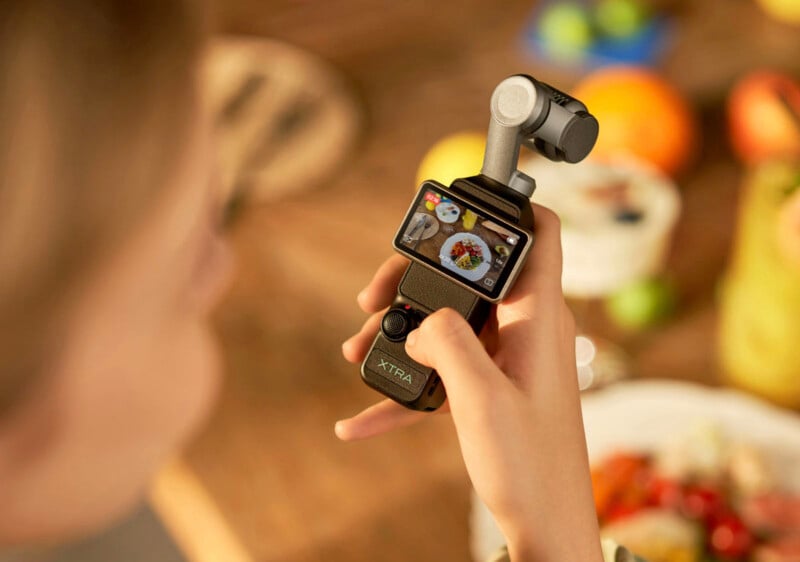 A person holds a small handheld camera with a screen displaying a close-up of a food dish, while other food items are blurred in the background on a wooden table.