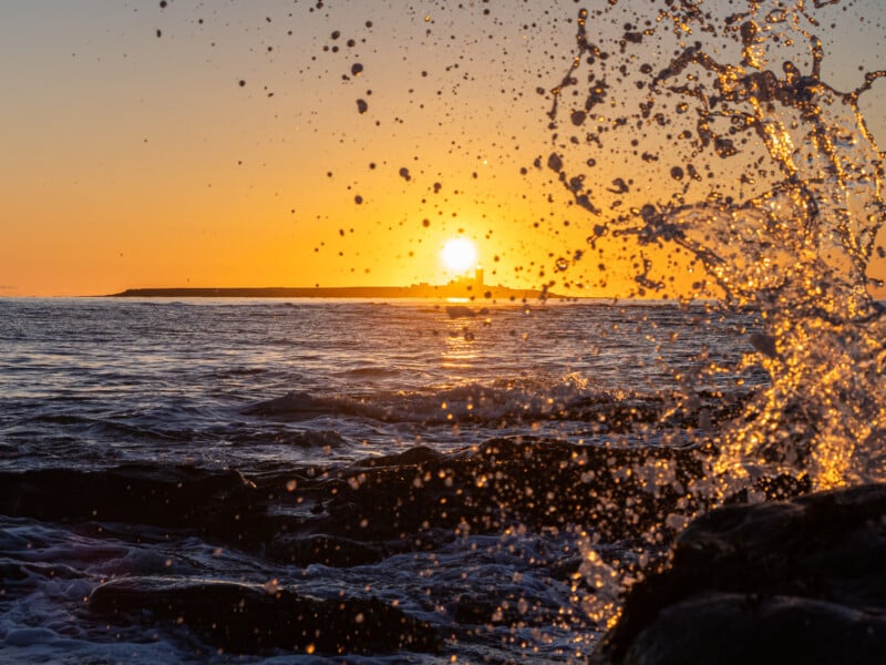Ocean waves splash against rocks in the foreground as the sun sets over the horizon, creating a vibrant orange glow across the sky and water.