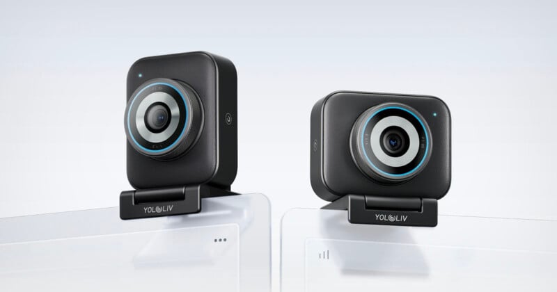 Dos cámaras web rectangulares negras con detalles circulares en azul están montadas sobre un monitor de computadora blanco. marca "Yulolifu" Visible en la base de cada cámara web. El fondo es sencillo y claro.