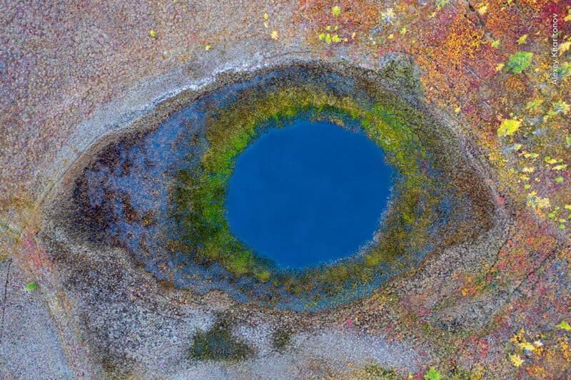 Aerial view of a circular blue pond surrounded by green, brown, and yellow vegetation, resembling an eye shape in the landscape.