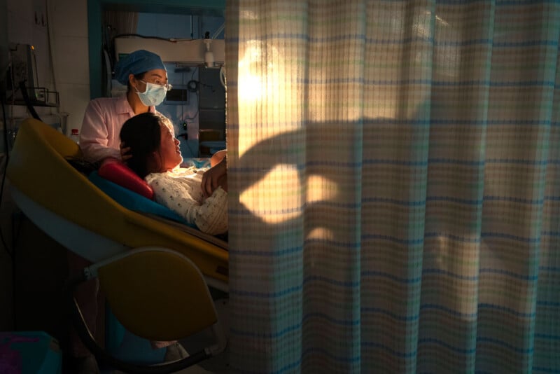 A nurse in scrubs and a mask supports a woman in labor lying on a hospital bed. Warm light shines through a striped privacy curtain, casting soft shadows in the delivery room.