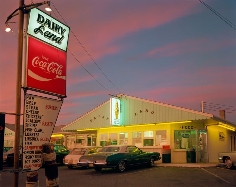 A retro Dairy Land diner glows with yellow lights at dusk, featuring a large Coca-Cola sign and a menu board. Classic cars are parked outside under a pink and blue sky.