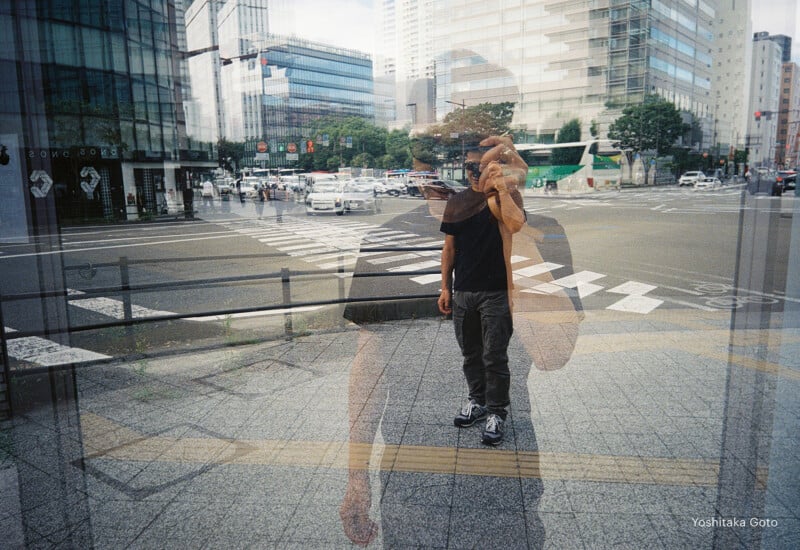 A person taking a photo is reflected in a glass window, blending with the busy city street, tall buildings, and traffic visible behind them. The effect creates a layered, double-exposure-like scene.