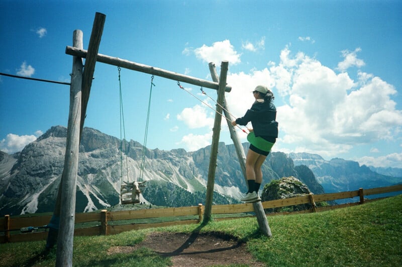 A person in a cap and jacket stands on a wooden swing structure set in a grassy field, with dramatic mountain peaks and a partly cloudy sky in the background.