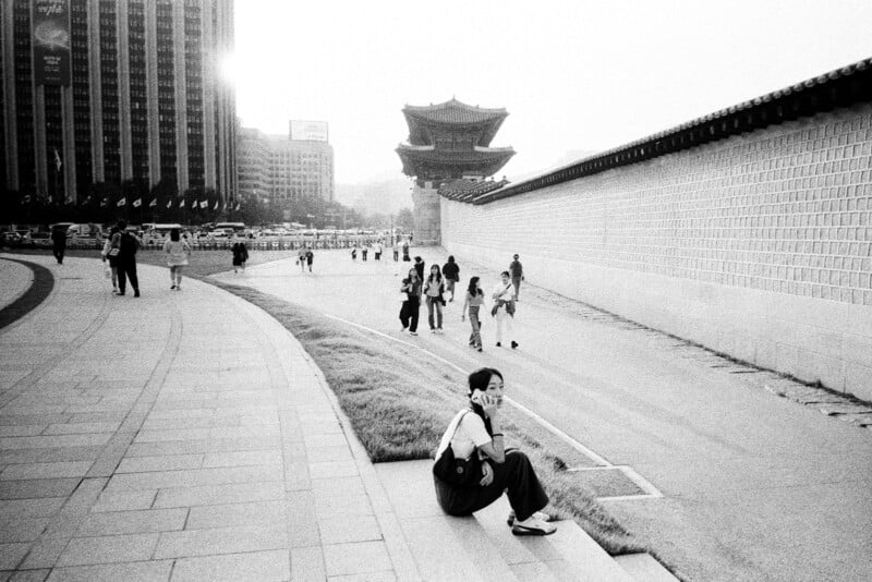 A woman sits on steps beside a large stone wall, talking on her phone. Several people walk along a wide path in the background, leading toward a traditional Korean gate. The scene is bright and urban, with tall buildings nearby.
