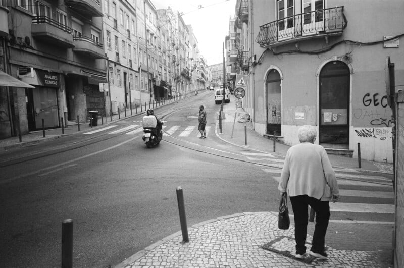 A black and white photo of a city street shows an elderly woman walking on a sidewalk, a scooter with two riders driving away, and a person crossing the street on a crosswalk. Buildings line both sides of the uphill street.