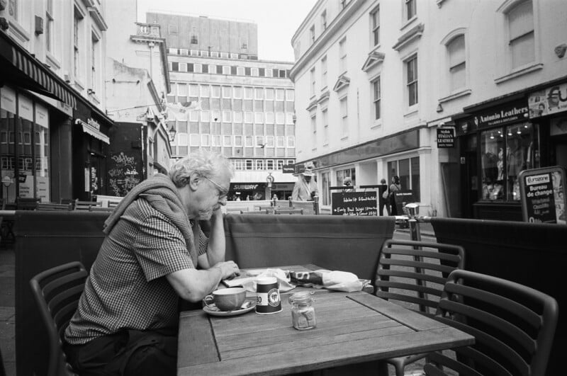 An older person sits alone at an outdoor café table, head resting on hand, with coffee cups and plates in front. The street is lined with shops and empty chairs, and tall buildings are visible in the background.