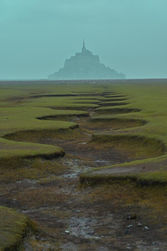 Un paisaje brumoso con hierba agrietada que conduce a un monasterio isleño en la distancia, recortado contra un cielo azul pálido. La escena da a la gente un sentimiento de paz y misterio.