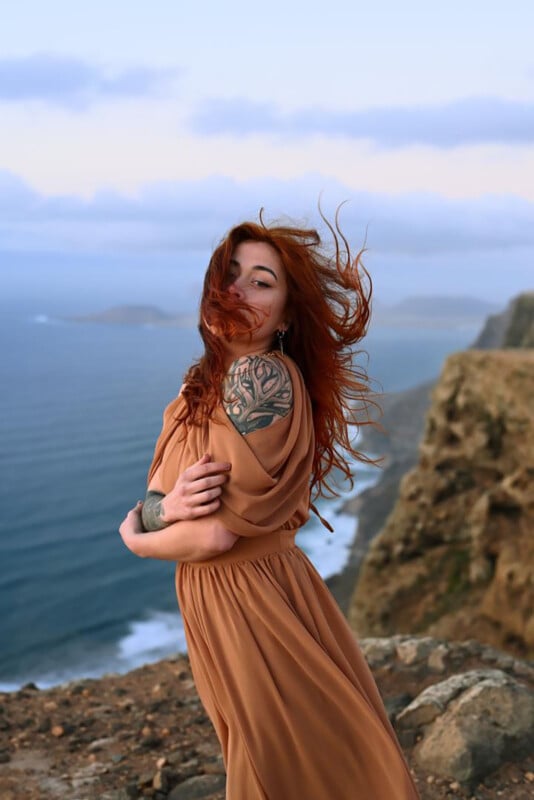 Una mujer de largo cabello rojo y vestido con un vestido color canela se encuentra al borde de un acantilado rocoso junto al mar, con el cabello ondeando al viento. Tiene un gran tatuaje floral en el hombro y mira con confianza a la cámara.