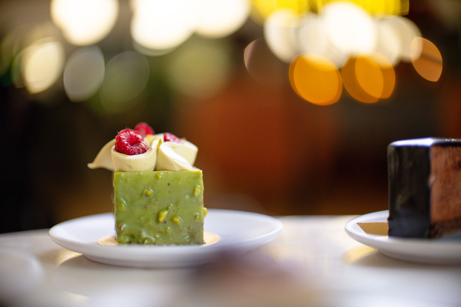 Un pequeño pastel cuadrado de pistacho cubierto con frambuesas y chocolate blanco en un plato blanco con otro postre difuminado en el fondo y luces bokeh.