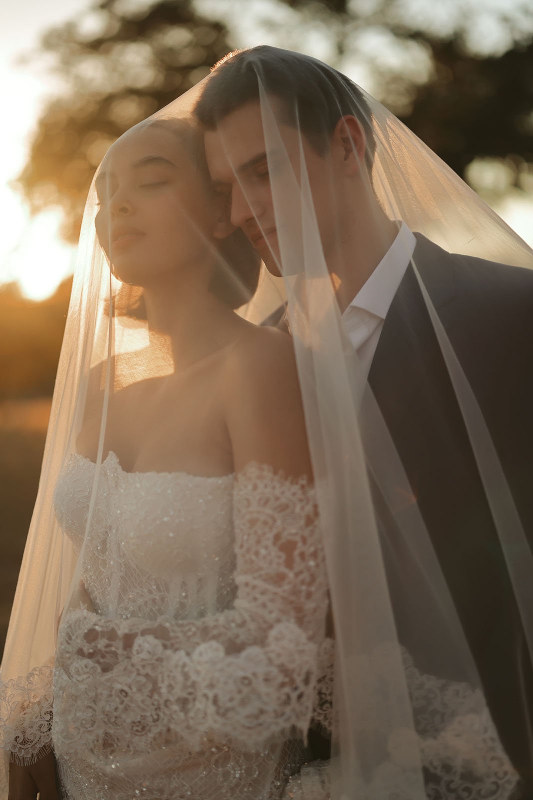 Al atardecer, los novios, envueltos en velos transparentes, permanecían al aire libre. La novia, con un vestido palabra de honor de encaje, tiene los ojos cerrados y el novio inclina suavemente la cabeza hacia ella.