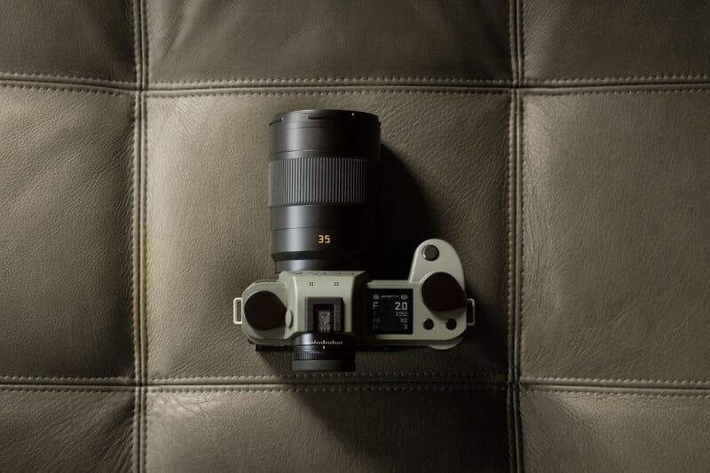 A digital camera with a large 35mm lens rests on a textured, gray leather surface, viewed from above. The camera’s display screen and controls are clearly visible.