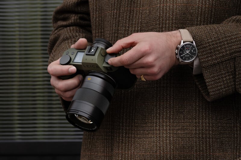 A person wearing a brown plaid coat and a wristwatch adjusts settings on a Leica camera, holding it with both hands in front of a striped, blurred background.