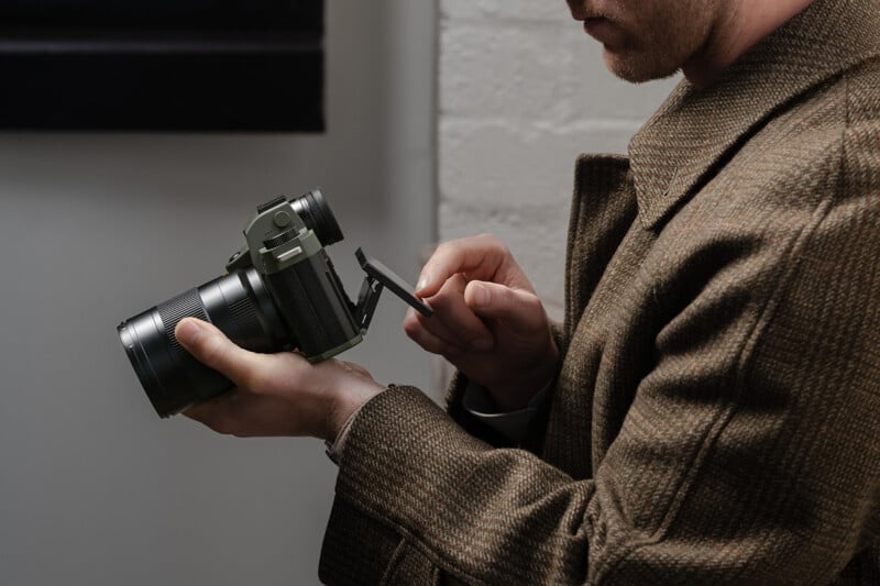 A person wearing a brown coat holds a digital camera in one hand and adjusts its flip-out screen with the other, standing indoors near a white wall.