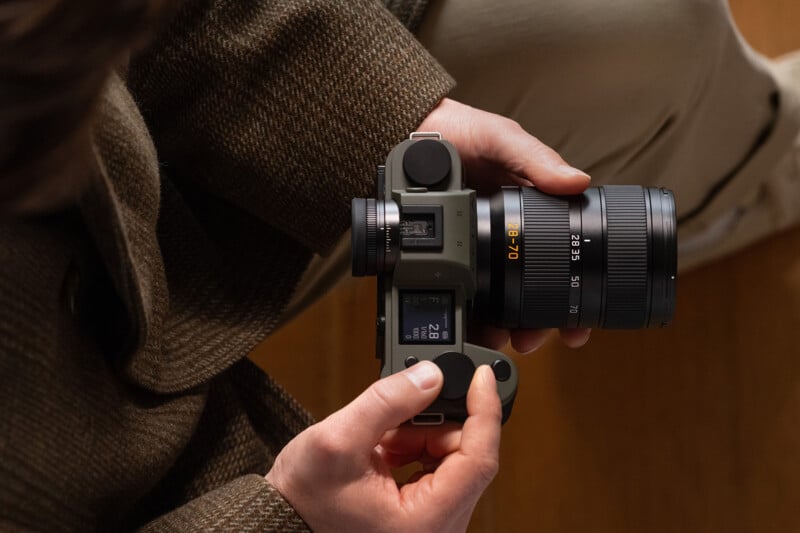 A person in a brown coat holds a digital camera with a 28-70mm lens, adjusting the settings on the top display screen. The image is shot from above, focusing on the camera and the person’s hands.
