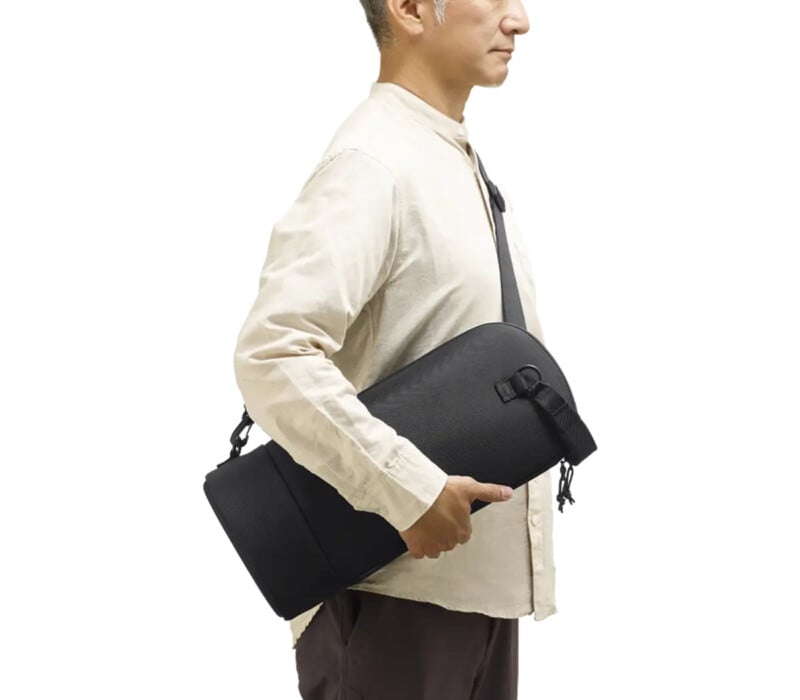 A person in a light beige shirt carries a black yoga mat bag with a shoulder strap, holding it under one arm while standing in profile against a plain background.