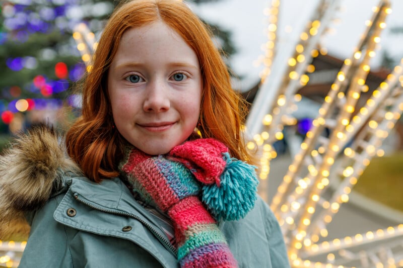 Una joven pelirroja y una colorida bufanda sonríe frente a las luces navideñas al aire libre. Llevaba un abrigo verde con capucha forrada de piel. El fondo está desdibujado por luces y árboles.