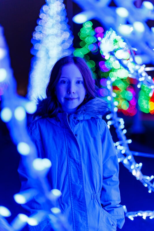 Un niño que lleva un abrigo de invierno azul claro se encuentra entre brillantes y coloridas luces navideñas que brillan en azul y violeta, rodeado de decoraciones borrosas y árboles brillantes en el fondo.