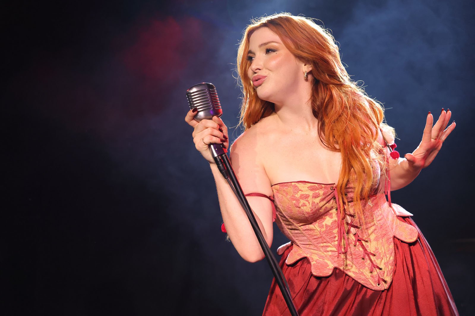 Una mujer con el pelo largo y rojo que lleva un corsé rojo y dorado se alza sobre un fondo oscuro y ahumado y canta en un micrófono antiguo.