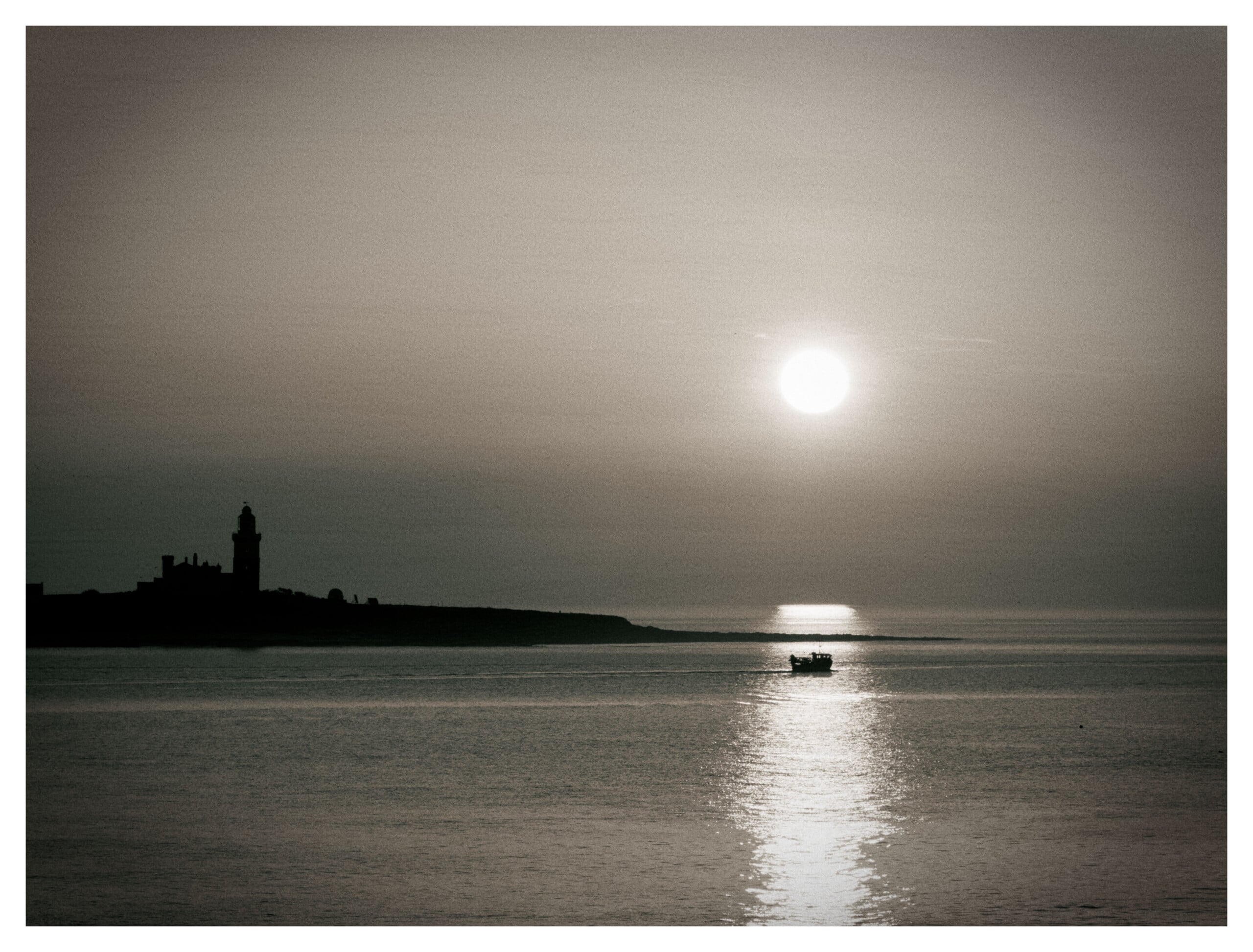 Al atardecer, un pequeño barco flota sobre las tranquilas aguas, con el sol bajo en el cielo y reflejándose en el mar. Al fondo se ve la costa lejana y un faro. La imagen tiene un tono sepia.