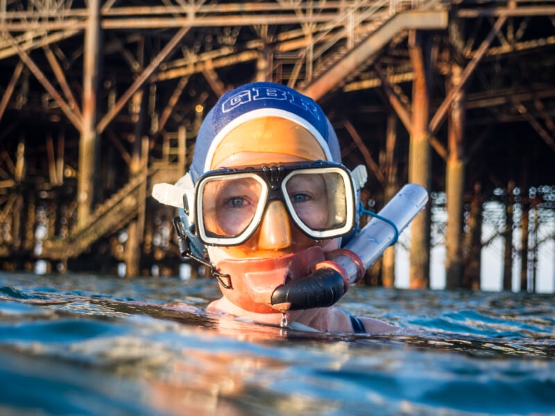 Un hombre que lleva una máscara de snorkel y un gorro de baño flota en el agua con solo la cabeza fuera del agua. Al fondo se aprecia la estructura de un gran muelle o muelle.