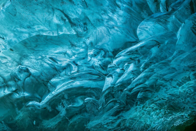 Close-up view of textured blue ice, showing swirling patterns and smooth, translucent surfaces with various shades of blue and white, resembling an icy cave or glacier wall.