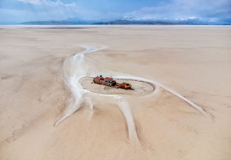 Una vista de pájaro de un edificio abandonado y vehículos oxidados, rodeado por un desierto seco y agrietado con líneas de sal blanca que irradian hacia afuera y montañas en la distancia bajo un cielo nublado.