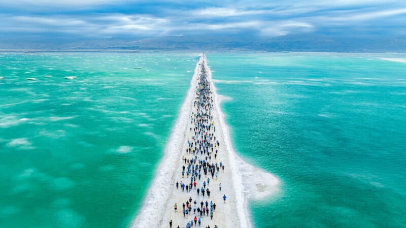 Un nutrido grupo de personas caminaba por un estrecho sendero blanco, bajo un cielo nublado y rodeado de agua turquesa, que creaba un marcado contraste con el mar.