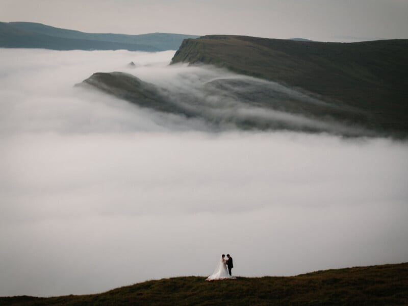 Una pareja parada en una montaña con nubes al fondo.