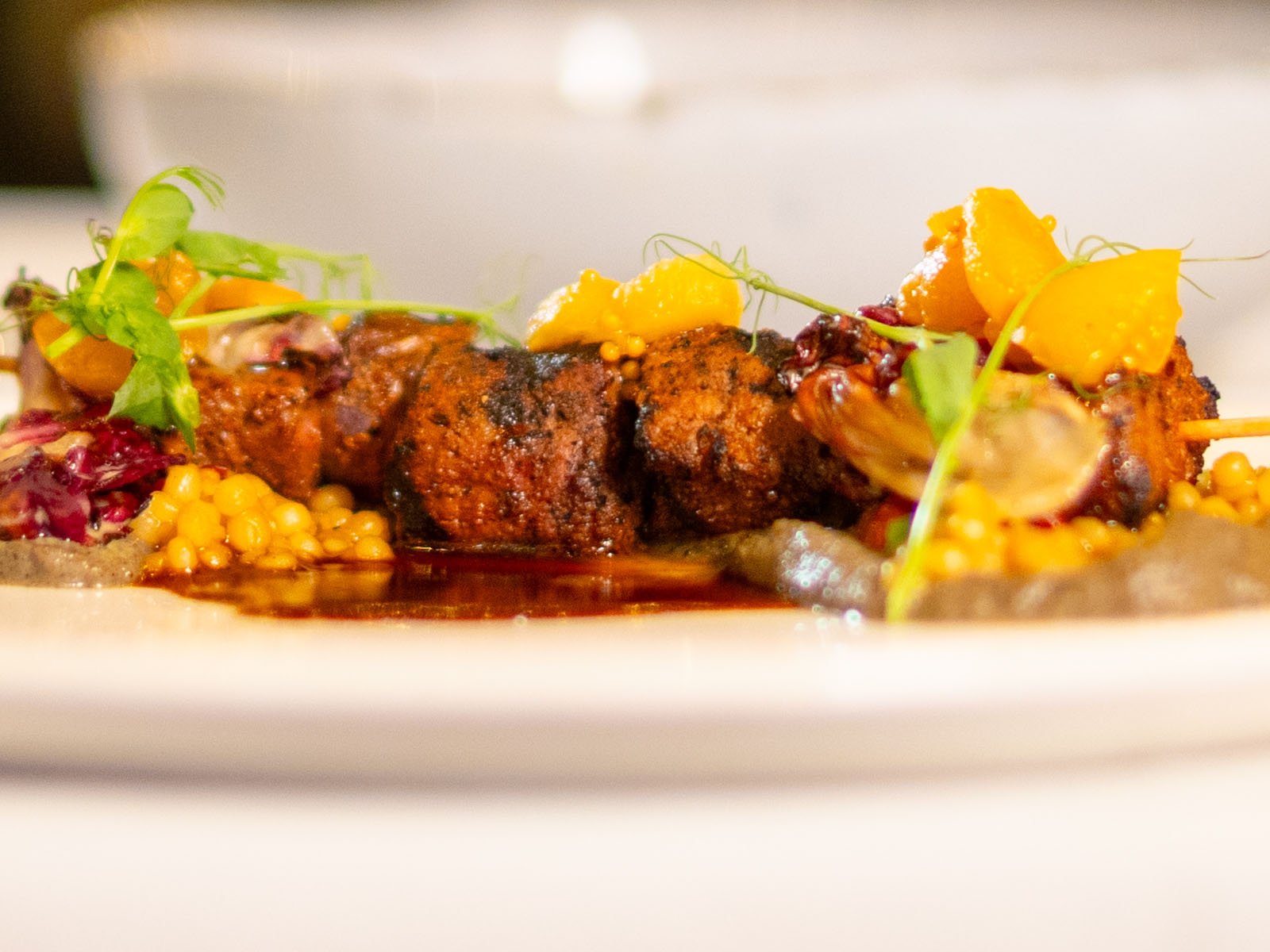 Primer plano de deliciosos kebabs cubiertos con carne sazonada a la parrilla sobre una cama de cuscús perlado de color amarillo brillante, adornado con microvegetales y vegetales amarillos cortados en cubitos, en un plato blanco.