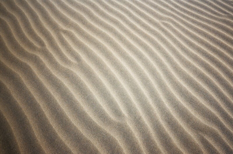 Wavy lines of wind-swept sand create a pattern of gentle, parallel ripples on the surface of a beach or desert. The texture highlights the fine grains and natural movement of the sand.