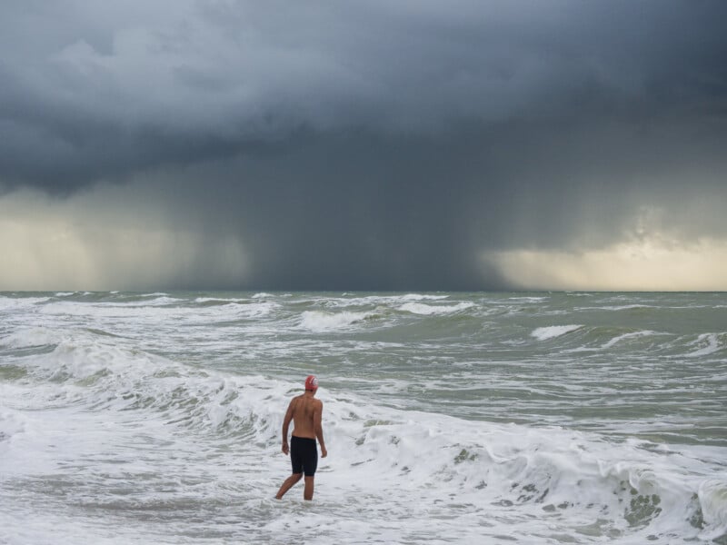 Un hombre con gorro de baño estaba de pie al borde del mar embravecido, frente a las furiosas olas. Bajo el cielo oscuro, hacía viento y llovía, y caía una fuerte lluvia a lo lejos.
