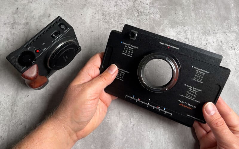 A person holds a black lens adapter labeled "45SL L-Mount" in their right hand, with a compact camera featuring a brown grip resting on a gray textured surface nearby.