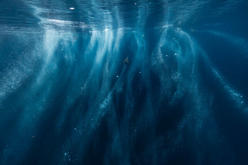Un buceador solitario nada bajo el agua, rodeado de rayos de luz y burbujas ascendentes en el mar azul profundo.