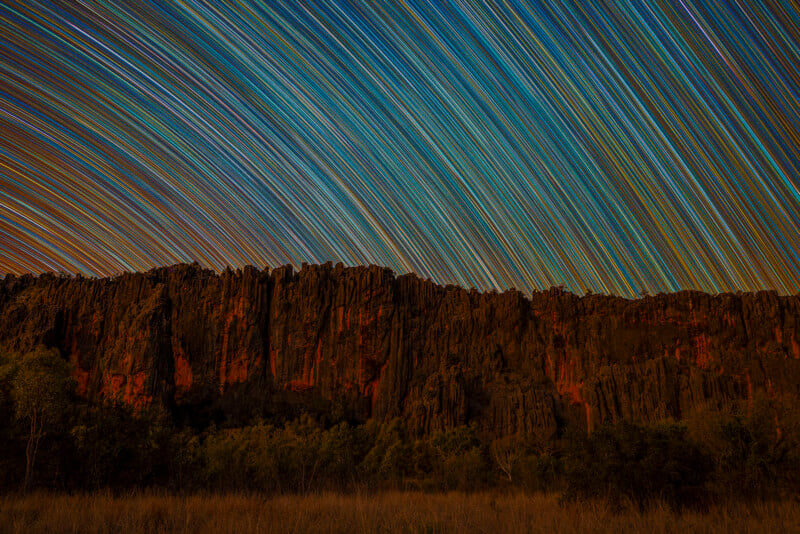 Coloridos senderos de estrellas atraviesan el cielo nocturno sobre formaciones rocosas oscuras y escarpadas y escasa vegetación, creando una escena vibrante y dinámica de movimiento celeste.