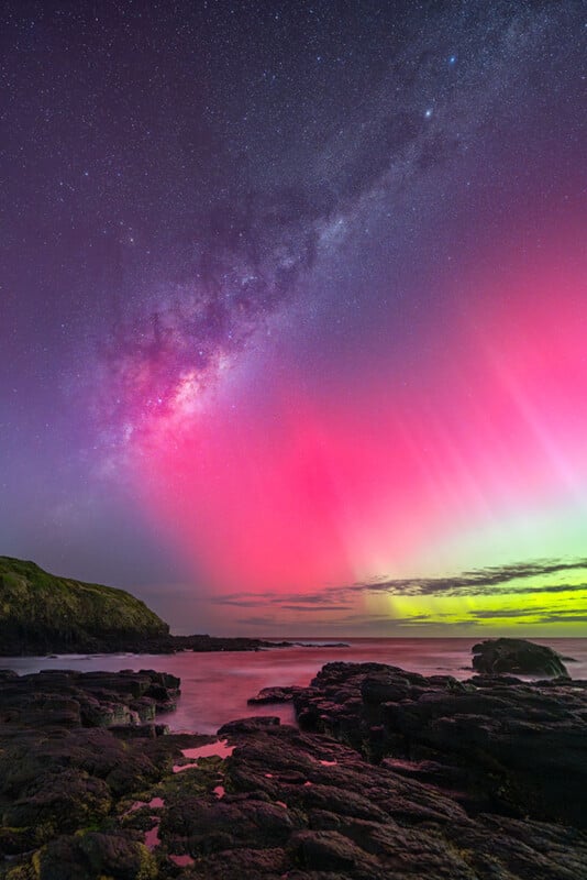 El vibrante cielo nocturno muestra la Vía Láctea, con auroras rosadas y verdes brillando sobre costas rocosas y aguas tranquilas. A la izquierda se ve una colina cubierta de hierba bajo un cielo estrellado y colorido.