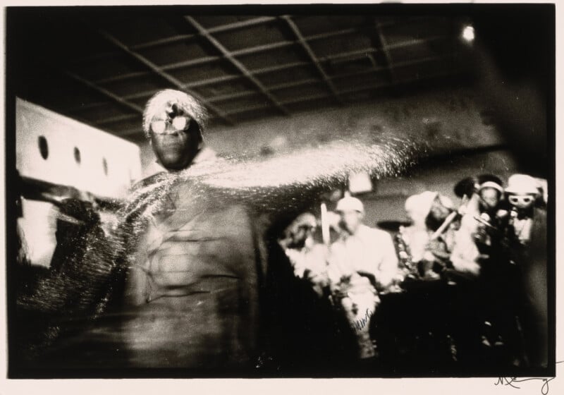 Una figura con un traje brillante y un tocado baila enérgicamente alrededor del escenario en un movimiento borroso. Al fondo, un grupo de músicos toca instrumentos en una habitación con poca luz.