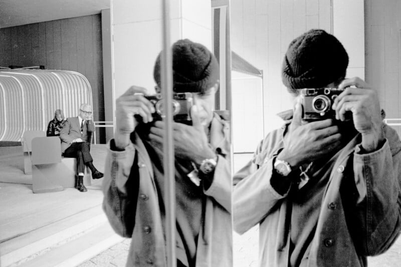 Hombre con gorro tomando fotos con una cámara frente a una superficie reflectante, creando una imagen reflejada. Al fondo, dos personas sentadas en un banco dentro de un edificio moderno.