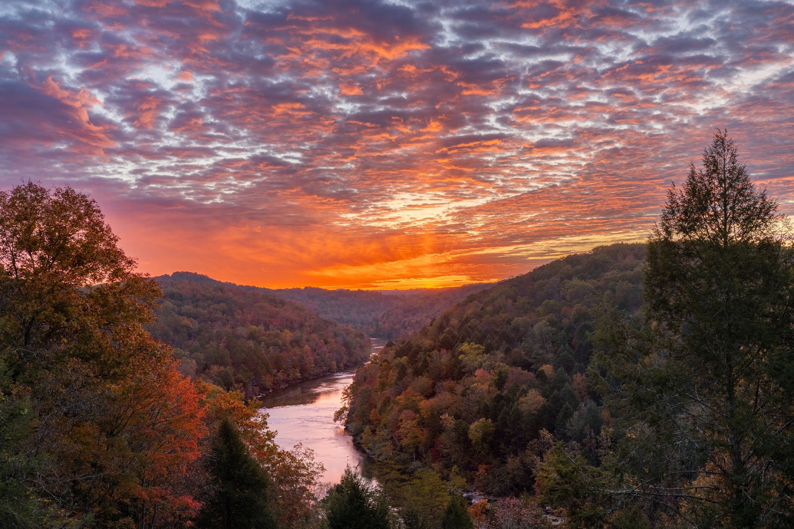 Una vibrante puesta de sol ilumina un río que serpentea a través de un valle boscoso, el cielo se llena de nubes coloridas y los árboles otoñales cubren las colinas a ambos lados del agua.