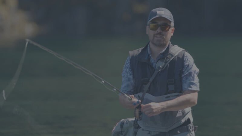 A man wearing sunglasses, a cap, and a fishing vest stands outdoors while fly fishing, holding a fishing rod over green water.