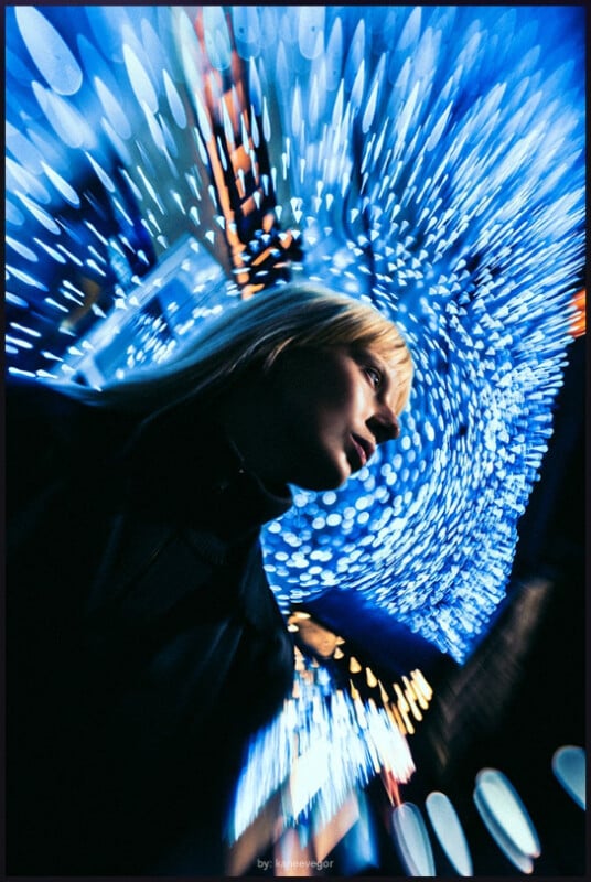 A person with blond hair stands under bright, swirling blue lights at night, creating a dynamic, futuristic atmosphere. The background is filled with glowing bokeh, giving a sense of motion and energy.