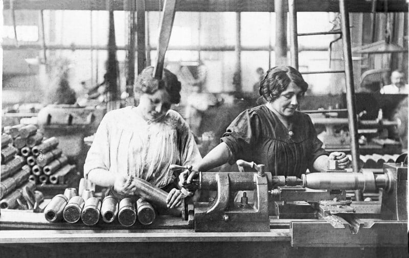 Two women work in a factory assembling artillery shells during World War I. They wear blouses and focus on handling metal shells at a workbench surrounded by tools and equipment.