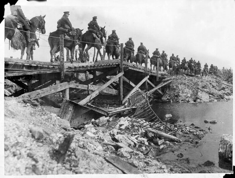 A line of soldiers on horseback crosses a damaged wooden bridge over rubble and water in a war-torn landscape, under a cloudy sky.