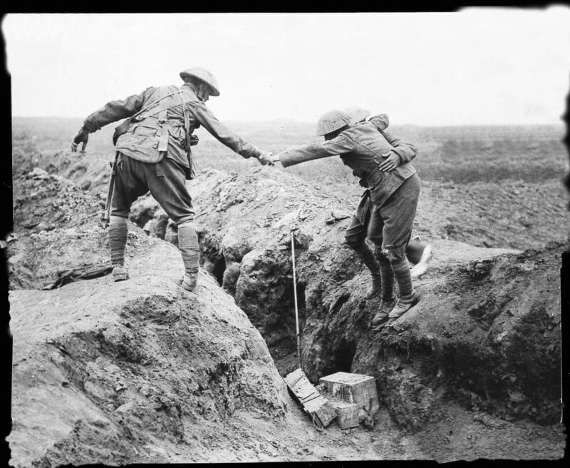 A soldier in uniform helps another soldier climb out of a muddy trench on a battlefield. The landscape is barren and desolate, with minimal vegetation and cloudy skies.