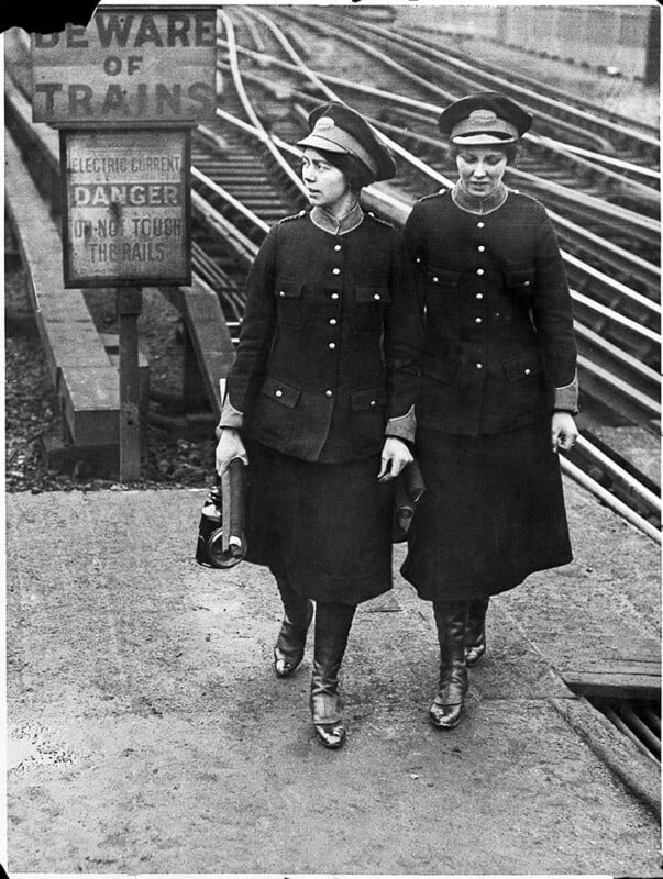 Two women in uniform walk side by side near train tracks. Both wear hats, jackets, and skirts. A sign nearby warns of electric danger and trains. The scene appears historical, possibly from the early 20th century.
