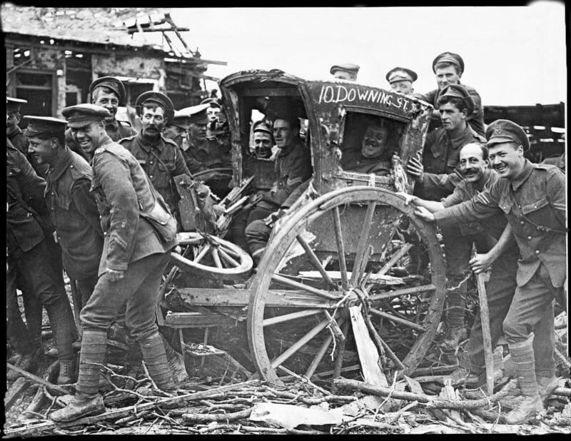 A group of soldiers in uniform pose cheerfully around a damaged, old carriage labeled "10 Downing Street" amid debris, suggesting a moment of levity during a wartime scene.