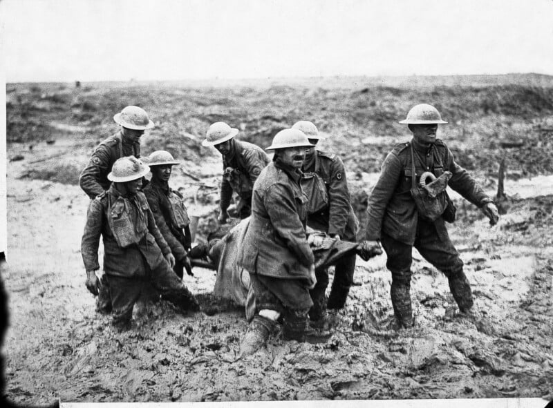 Six World War I soldiers in muddy uniforms and helmets carry a wounded comrade on a stretcher across a muddy, barren battlefield; the sky is overcast and the scene appears grim and bleak.